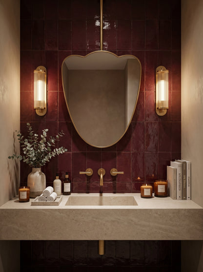 Luxury ceiling-mounted brass mirror with sculptural silhouette against glossy burgundy tiles and brass fixtures.