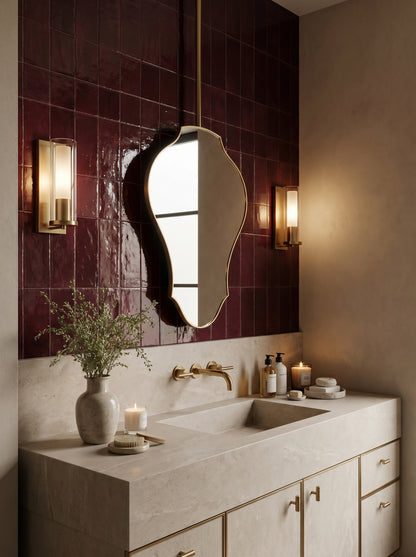 Sculptural brass mirror mounted above a stone vanity with burgundy tiled walls.