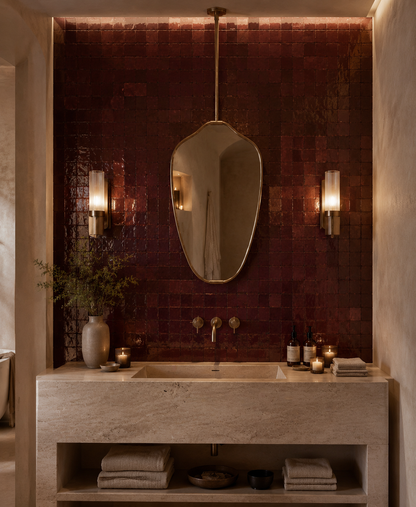 A luxury bathroom vanity featuring a textured, dark reddish-brown mosaic tile backsplash, a large organic-shaped wall mirror, and a stone sink console with folded towels and candles.