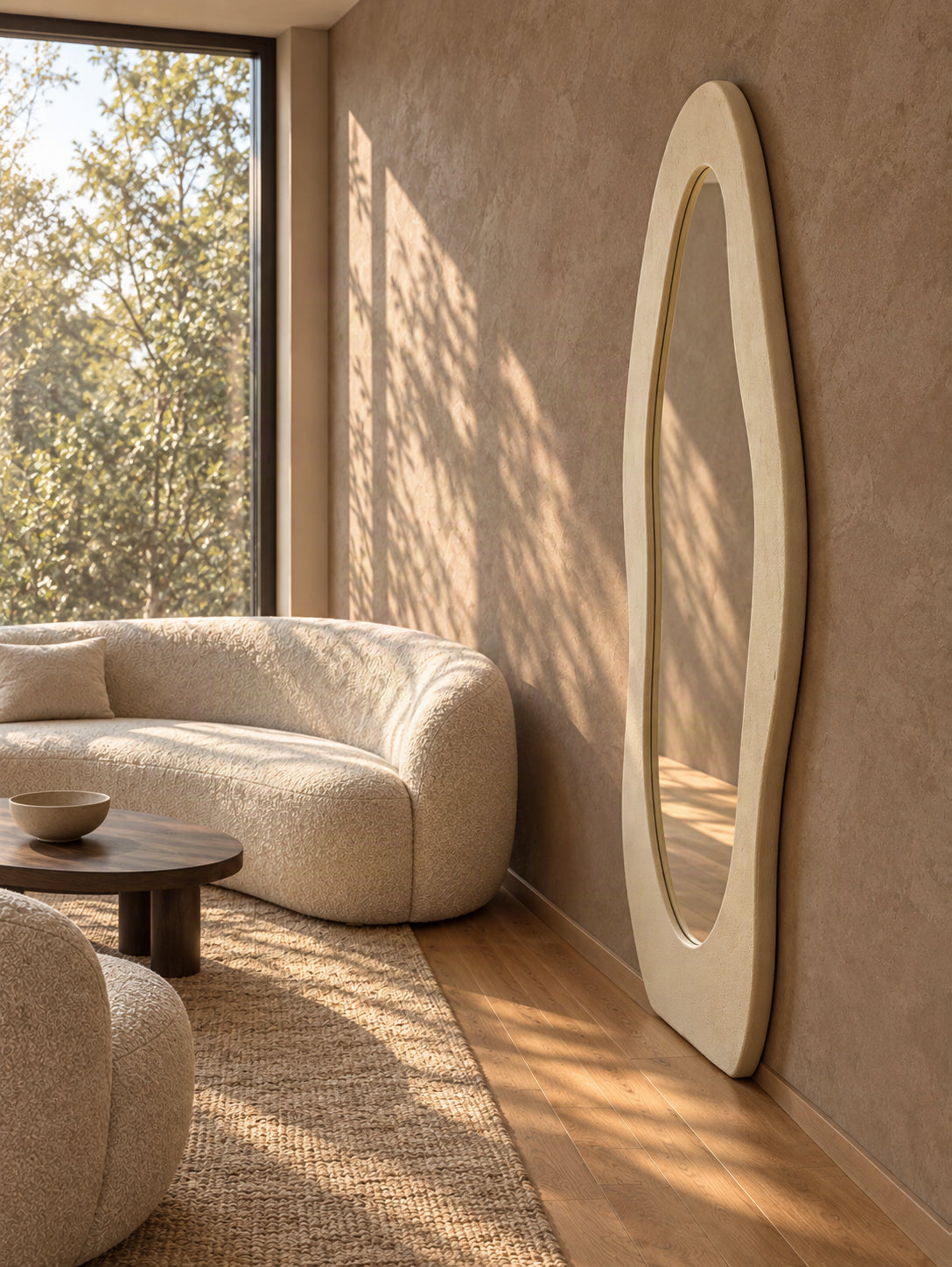 An organic-shaped cream mirror leaning against a textured beige wall in a sunlit modern living room with a curved cream boucle sofa and wood flooring.