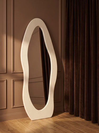 An organic-shaped floor mirror with a thick white frame leans against a brown paneled wall on a wooden floor next to dark brown curtains.