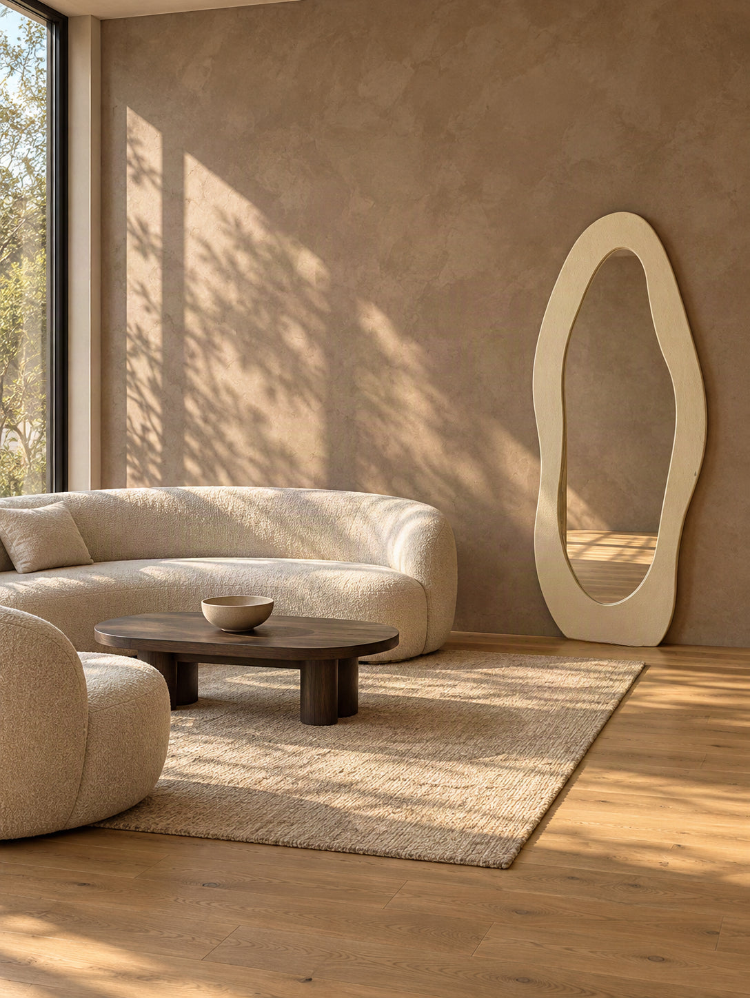 A modern living room scene with a curved cream boucle sofa, matching armchair, dark wood coffee table, textured rug, and a tall organic-shaped cream floor mirror against a warm stucco wall.