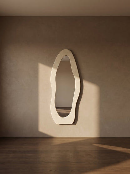 An organic-shaped cream wall mirror with a thick, wavy frame, mounted on a textured beige wall in a room with warm sunlight and a dark wood floor.