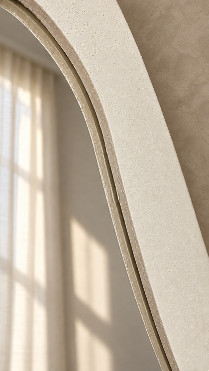 Close-up of a large arched mirror with a textured cream-beige frame, reflecting soft sunlight and window shadows against a warm neutral wall.
