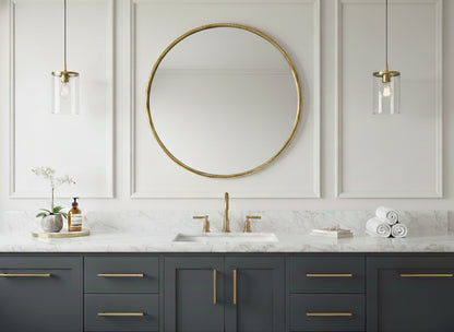Round brass mirror over a gray floating vanity on a marble bathroom wall.
