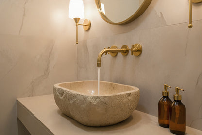 Natural stone vessel sink with wall-mounted brass faucet and marble wall.