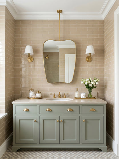Suspended wavy brass mirror above a sage green vanity with beige subway tiles and brass wall sconces in a traditional bathroom.