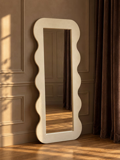 A tall, full-length floor mirror with a wavy scalloped frame, leaning against a brown paneled wall on a wooden floor next to dark brown curtains.