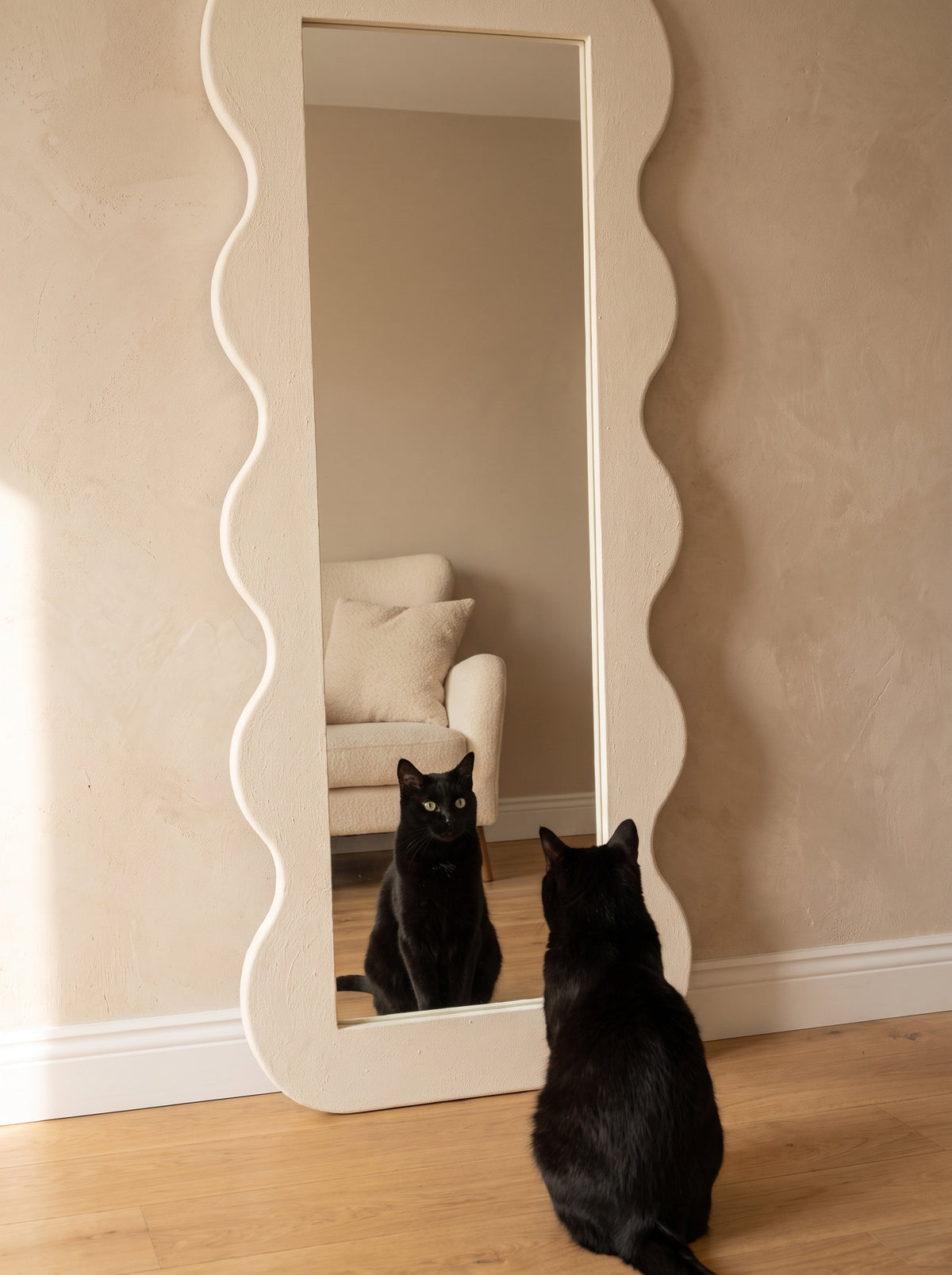 A tall floor mirror with a wavy scalloped frame stands against a beige wall on a light wood floor. A black cat sits in front of the mirror, looking at its reflection, which shows a cream armchair in the room.