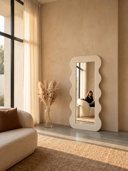 A tall, floor-length standing mirror with a wavy scalloped white frame leans against a beige plaster wall in a sunlit modern room. The mirror reflects a person sitting in a chair, while the room itself features a cream sofa, a vase of pampas grass, and a woven rug.