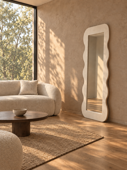 A tall, full-length floor mirror with a wavy, scalloped white border stands in a sunlit living room next to a beige textured sofa and a round wooden coffee table.