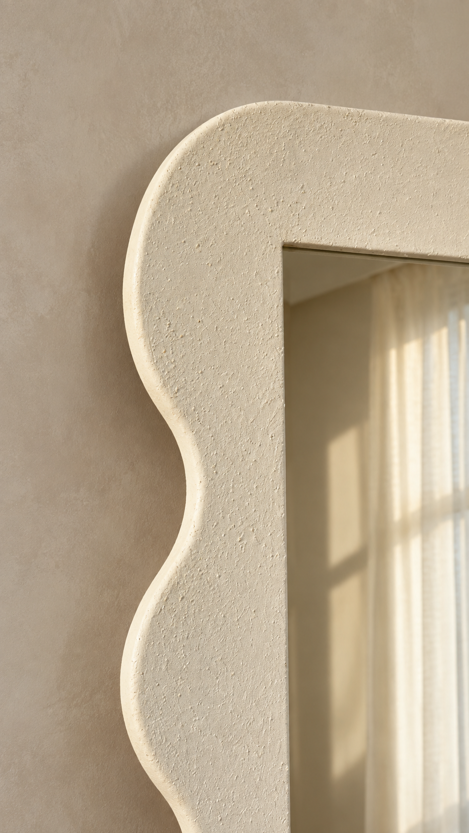 A close-up of a textured cream-colored wall mirror with a decorative scalloped border, mounted on a matching beige plaster wall with soft sunlight filtering through a curtain.