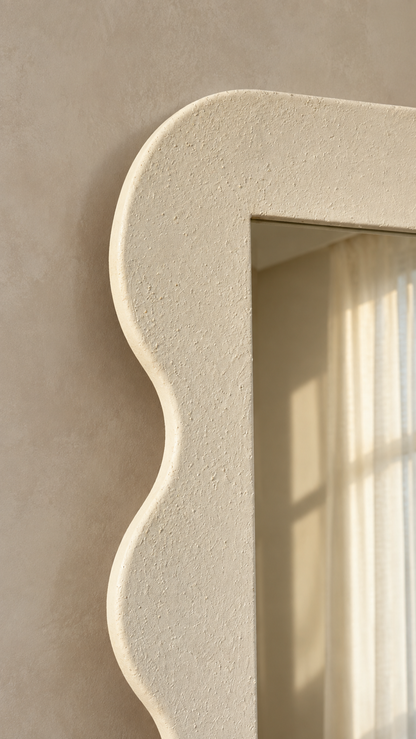 A close-up of a textured cream-colored wall mirror with a decorative scalloped border, mounted on a matching beige plaster wall with soft sunlight filtering through a curtain.