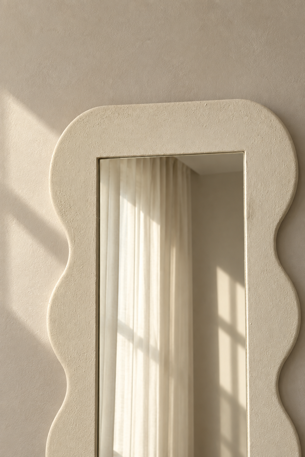 A textured cream-colored wall mirror with a wavy scalloped border, mounted on a matching beige plaster wall. Soft sunlight streams across the surface, reflecting a neutral-toned room with a curtain.