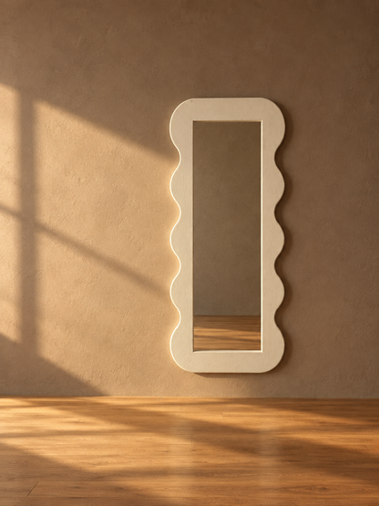 A tall, narrow rectangular wall mirror with a wavy scalloped border, shown leaning against a textured beige wall on a wooden floor with warm sunlight and shadows.