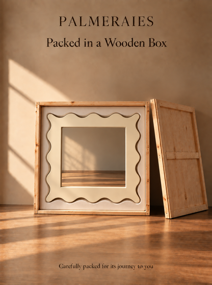 A wooden presentation box with a decorative wavy-edged window panel, shown open with its back panel leaning against a warm beige wall on a wooden floor. Text at the top reads 'PALMERAIES Packed in a Wooden Box' and smaller text at the bottom says 'Carefully packed for its journey to you'.